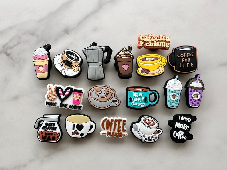 Op de afbeelding: Een verzameling van 15 kleurrijke schoenbedels met een koffie-thema. De bedels tonen verschillende koffiedrankjes, koffiezetapparaten en koffiegerelateerde zinnen zoals "Coffee is Please..." en "Need More Coffee".