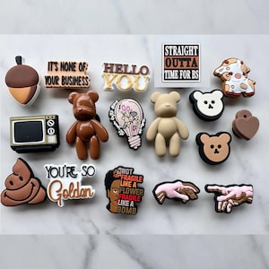 May include: A collection of 15 colorful shoe charms featuring various designs, including a cartoon bear, a lightbulb, a hand gesture, a poop emoji, a cow, an acorn, a television, a heart, and text that reads "Hello You", "It's None of Your Business", "You're So Golden", "Straight Outta Time For BS", and "Not Fragile Like A Flower Fragile Like A Bomb".