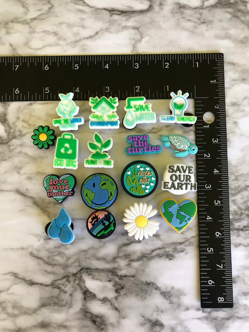 May include: A collection of 14 colorful shoe charms with various environmental themes. The charms feature images of flowers, turtles, hearts, water drops, a lightbulb, a house, a tree, and the Earth. The charms also include text such as "Save the Turtles", "Save Our Earth", "Love Your Mother", and "Friend to Bugs".