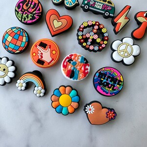 May include: A collection of 16 colourful, retro-themed shoe charms featuring peace signs, flowers, hearts, rainbows, and a groovy van with the text "Love Peace".