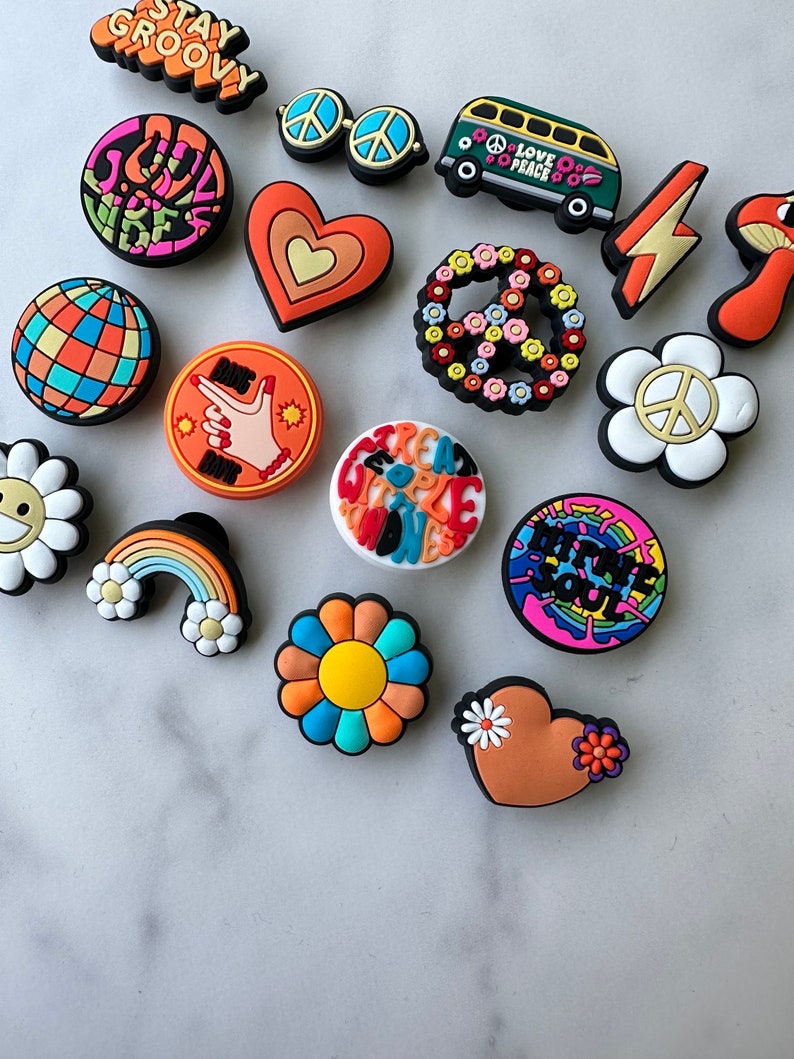 May include: A collection of 16 colourful, retro-themed shoe charms featuring peace signs, flowers, hearts, rainbows, and a groovy van with the text "Love Peace".