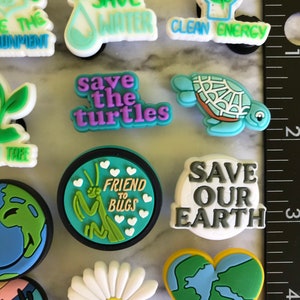 May include: A set of 11 colorful shoe charms with environmental themes. The charms feature images of a house plant, a water drop, a lightbulb, a turtle, a praying mantis, a daisy, a heart-shaped earth, and text that says "Save the Environment", "Save Water", "Clean Energy", "Save the Turtles", "Friend to Bugs", "Save Our Earth".