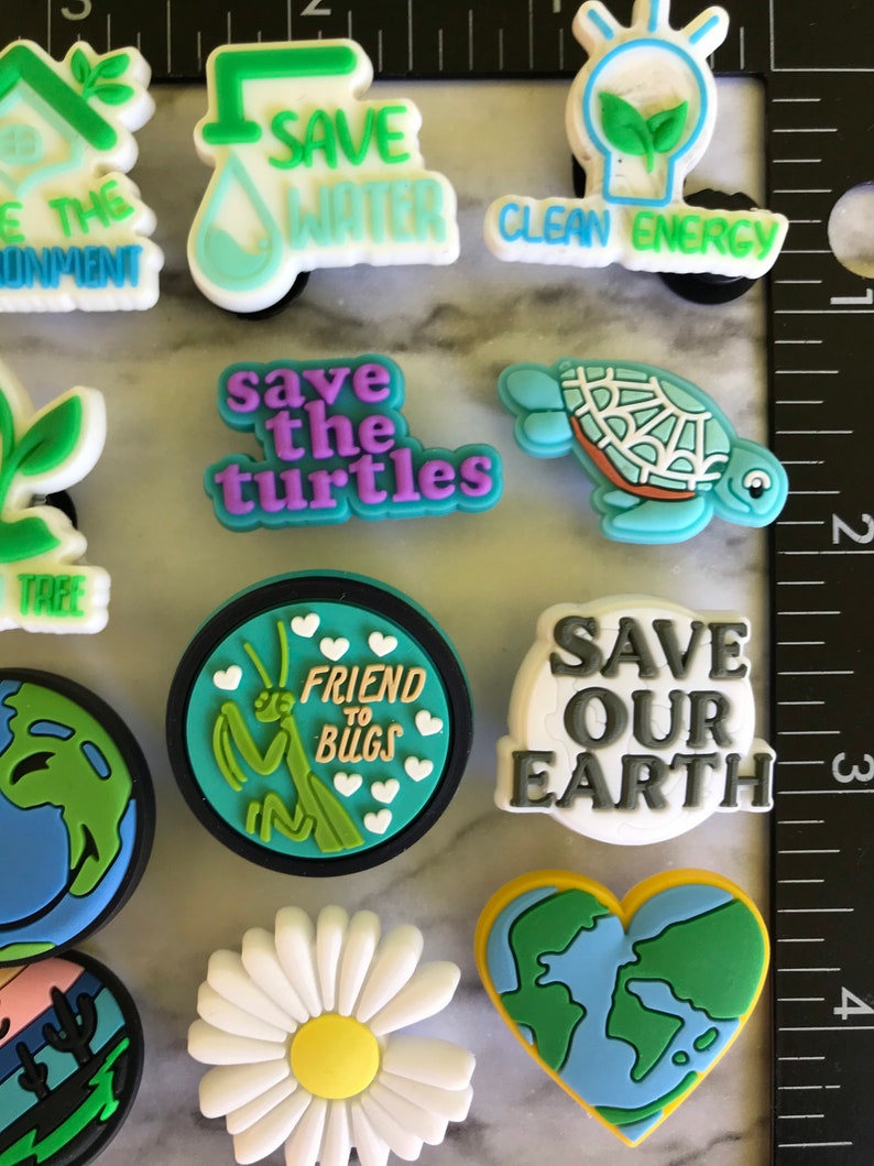 May include: A set of 11 colorful shoe charms with environmental themes. The charms feature images of a house plant, a water drop, a lightbulb, a turtle, a praying mantis, a daisy, a heart-shaped earth, and text that says "Save the Environment", "Save Water", "Clean Energy", "Save the Turtles", "Friend to Bugs", "Save Our Earth".