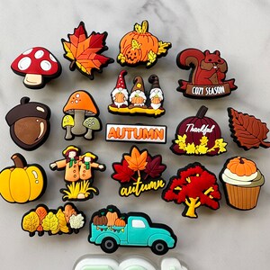 Op de afbeelding: Een verzameling van 17 kleurrijke herfst-schoencharms, waaronder pompoenen, bladeren, paddenstoelen, een eekhoorn, een kabouter, een vogelverschrikker, een vrachtwagen en een cupcake. De charms zijn gemaakt van siliconen en hebben een verscheidenheid aan herfstkleuren, waaronder oranje, rood, geel, bruin en groen.