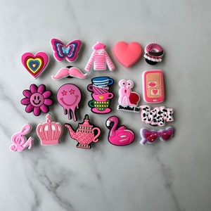 May include: A collection of 17 pink and white shoe charms with various designs, including a heart, a butterfly, a mustache, a teacup, a snail, a phone, a crown, a teapot, a flamingo, and sunglasses.