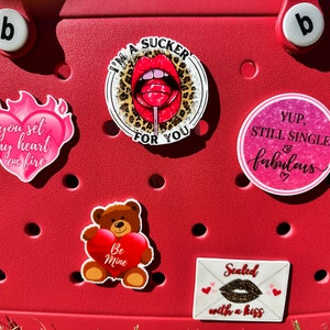 May include: A red tote bag with various Valentine's Day themed stickers. The stickers include a heart with flames, a teddy bear holding a heart, a red lip with a lollipop, a pink circle with the text "YUP, STILL SINGLE & fabulous", and a white envelope with the text "Sealed with a kiss".