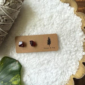Peut inclure: Deux petites boucles d'oreilles clous en grenat rouge brut sur une carte marron avec un logo d'arbre et le texte "forest & fog". Les boucles d'oreilles sont présentées sur une surface blanche texturée, avec un paquet de sauge et un plat doré décoratif en arrière-plan.