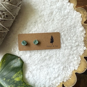 May include: Two small, square, green gemstone stud earrings presented on a brown card with the text "forest & fog" and a tree graphic. The earrings are displayed on a white, textured surface with a gold-coloured edge.