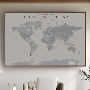 May include: A rectangular wall map featuring a grey world map design with the text "OUR ADVENTURES" and "CHRIS & SELENA" printed at the top. The map is framed in a dark wood border and displays continents in a darker grey against a light grey background.