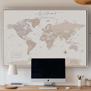 May include: A framed world map with a neutral color palette, featuring the text "The Williams Adventures Around the World." The map is displayed above a wooden desk with a computer monitor, keyboard, and lamp.