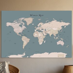 May include: A rectangular wall art print featuring a world map with light beige continents against a muted blue-grey background. The text "WORLD MAP" is printed in a dark font at the top centre, and the map displays detailed geographical labels.