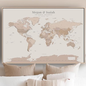 May include: A rectangular wall art print featuring a world map in shades of beige and tan. The text at the top reads Megan & Isaiah, Two Hearts, One World. The map displays continents and countries with a neutral colour palette and a thin black frame.