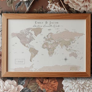 May include: A world map print in a light oak wood frame featuring the text Emily & Jacob Adventures Around the World. The map displays continents in muted beige and brown tones on a cream background with a compass rose and a legend.