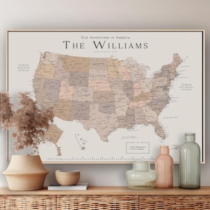 May include: A framed map of the United States with a neutral color palette. The map is labeled "Our Adventures in America" and "The Williams." Decorative vases and a woven basket sit on a wooden surface in front of the map.