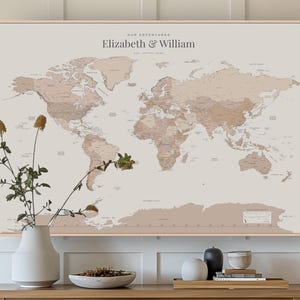 May include: A framed world map in beige and brown tones, featuring the text "Our Adventures Elizabeth & William". The map shows detailed country outlines. A white vase with dried flowers is in front of the map. The frame is light brown.