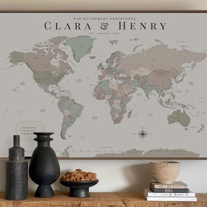 May include: A framed world map print with a muted colour palette featuring the text "OUR RETIREMENT ADVENTURES" and "CLARA & HENRY EST. 2026". The map displays continents in shades of sage green, dusty rose, and beige against a light grey background.