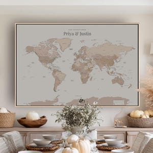 May include: A framed world map in shades of brown and beige, with the text "Our Adventures" and the names "Priya & Justin" at the top. The map is displayed on a light grey background, with a wooden frame.