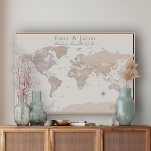 May include: A framed horizontal world map print featuring a light beige background with muted pink and brown continents. The text "EMILY & JACOB" and "Adventures Around the World" is printed in black at the top centre. A compass rose is visible in the lower right corner.