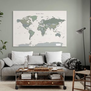 May include: A large world map canvas print in shades of green and gray hangs above a gray sofa. The map is labeled "World Map" and is mounted on a white background. A coffee table with books and a chess set sits in front of the sofa.