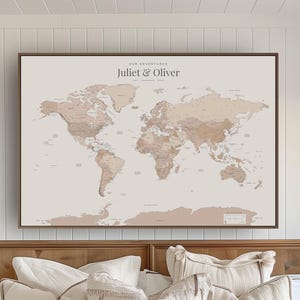 May include: A framed world map with a neutral color palette, featuring a brown wooden frame. The map displays continents and countries in shades of beige and brown. Text at the top reads "OUR ADVENTURES" and "Juliet & Oliver".