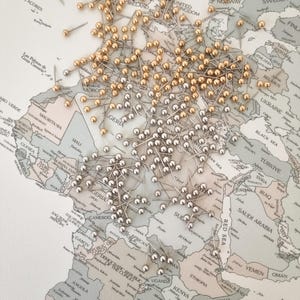 Peut inclure: Une carte du monde avec de nombreuses épingles dorées et argentées indiquant des lieux. La carte présente une palette de couleurs neutres avec des noms de pays. Les épingles sont regroupées dans diverses zones, suggérant des voyages ou de la planification.