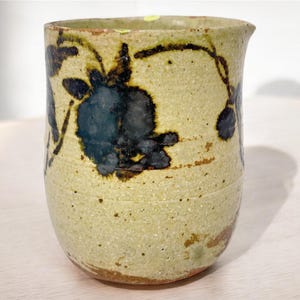 May include: A small, off-white ceramic jug with a brown and black floral design. The jug has a rounded body and a slightly flared lip.