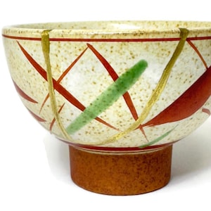 May include: A ceramic bowl with a speckled beige exterior, decorated with red and green leaf-like designs and gold accents. The bowl has a brown base and a rim with a red stripe. The bowl is likely used for serving food.