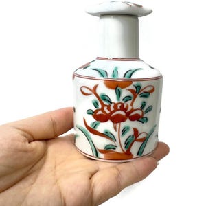 以下が含まれることがあります： 赤、緑、オレンジの花柄が施された白いセラミックボトル。ボトルは円筒形の本体と長い首を持ち、平らな丸いストッパーで覆われています。花柄はボトルの本体の周りに繰り返されています。