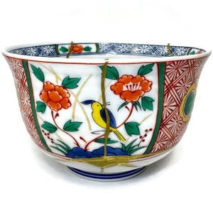 Könnte beinhalten: Eine weiße Keramikschale mit detailliertem Blumen- und Vogeldesign. Die Schale hat orangefarbene Blumen, grüne Blätter und einen gelben Vogel mit blauen Akzenten. Der Rand hat ein geometrisches Muster in Rot, Grün und Blau. Die Schale hat goldene Akzente und sichtbare Reparaturen.