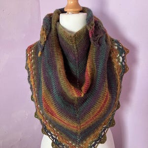 Könnte beinhalten: Ein handgestrickter Schal in Dreiecksform, auf einer Schaufensterpuppe präsentiert. Der Schal weist einen Farbverlauf auf, darunter Grün-, Braun-, Rot- und Violetttöne. Die Kanten sind mit einem gewellten Rand versehen.