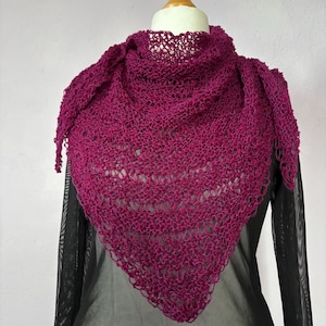 Könnte beinhalten: Ein handgestrickter Schal in Magenta, der auf einer Schaufensterpuppe präsentiert wird. Der Schal ist dreieckig, mit einem zarten Lochmuster und einem gewellten Rand. Der Strickstoff wirkt weich und fällt anmutig. Der Hintergrund ist eine hellrosa Wand.