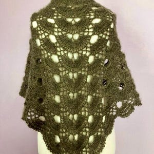 Könnte beinhalten: Ein dunkel olivgrüner gehäkelter Schal mit einem dekorativen Lochmuster. Der Schal wird auf einer Schaufensterpuppe präsentiert, die seine dreieckige Form und das aufwendige Design zeigt. Das Garn wirkt weich und strukturiert.