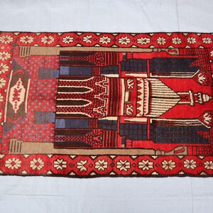 May include: A red, blue, and beige prayer rug with a mosque design. The rug has a fringe border and a floral pattern.