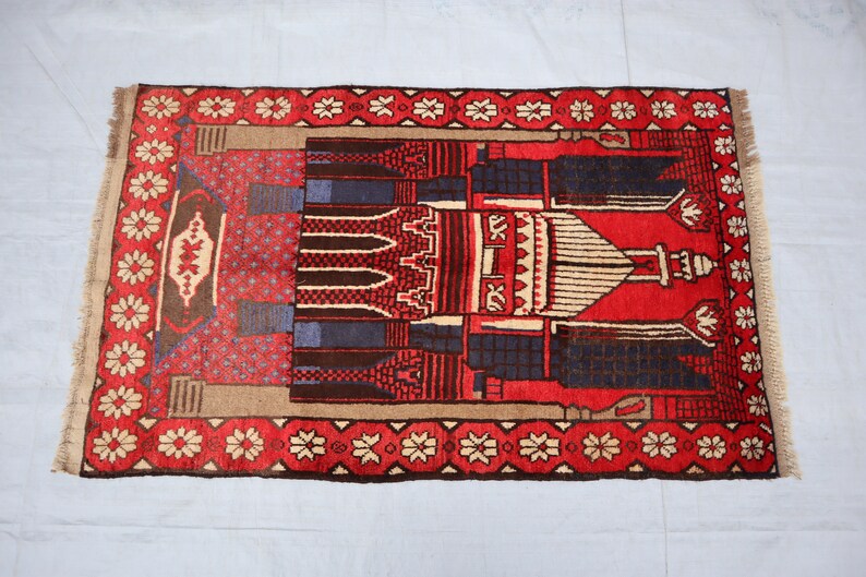 May include: A red, blue, and beige prayer rug with a mosque design. The rug has a fringe border and a floral pattern.