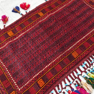 May include: A rectangular woven rug featuring a deep red and black geometric pattern with orange square motifs. The edges are adorned with long, braided red fringe tassels accented by colorful beads in blue, yellow, and pink hues along the border.