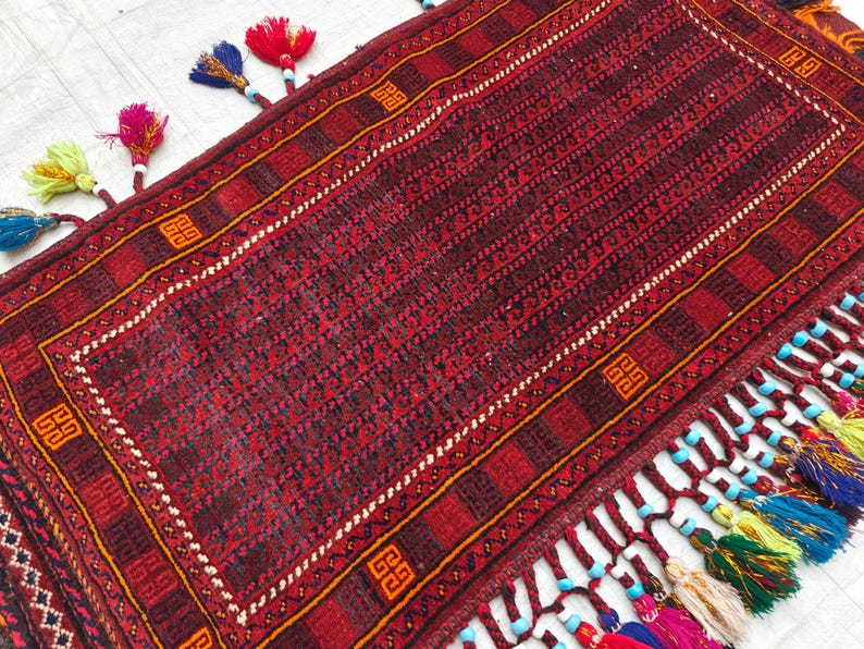May include: A rectangular woven rug featuring a deep red and black geometric pattern with orange square motifs. The edges are adorned with long, braided red fringe tassels accented by colorful beads in blue, yellow, and pink hues along the border.