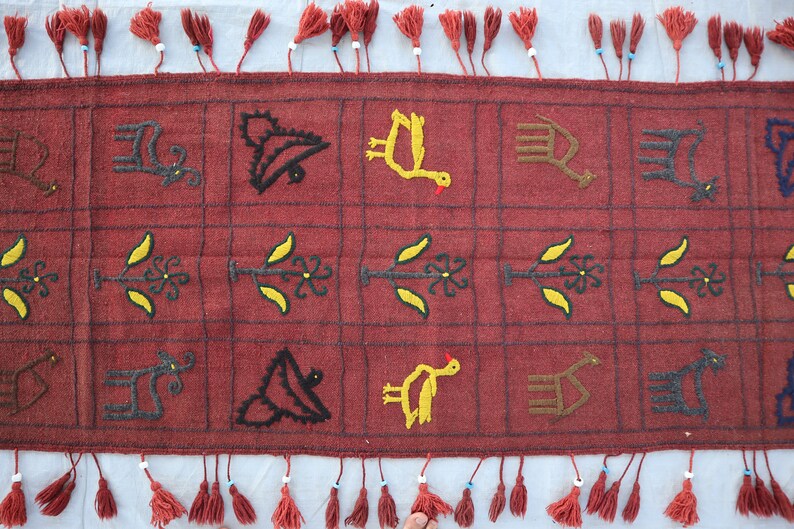May include: A red wool runner rug with embroidered designs of birds, plants, and animals. The rug has a fringe border with tassels.