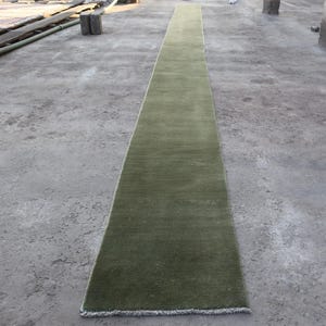 Könnte beinhalten: Ein langer, schmaler, olivgrüner Teppich. Der rechteckige Teppich hat ein schlichtes Design und Fransen an den kurzen Enden. Er liegt auf einer Betonoberfläche.