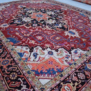May include: A traditional Persian rug with a red background and intricate floral and geometric patterns in shades of blue, cream, and black. The rug has a central medallion and a border with a repeating design.