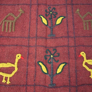 May include: A red woven tapestry with a grid pattern. The tapestry features embroidered images of camels, flowers, and birds. The birds are yellow with red beaks and the flowers are yellow with green leaves.