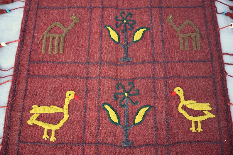 May include: A red woven tapestry with a grid pattern. The tapestry features embroidered images of camels, flowers, and birds. The birds are yellow with red beaks and the flowers are yellow with green leaves.