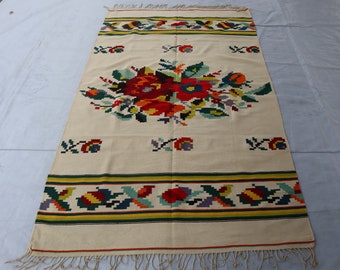 May include: A white woven rug with a colorful floral design in the center. The rug has a yellow, green, and red border.