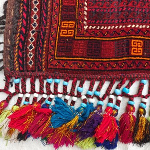 May include: A woven textile featuring a deep red background with intricate geometric patterns in black, orange, and white. The edge is finished with braided fringe adorned with small turquoise and white beads, ending in multi-colored yarn tassels.