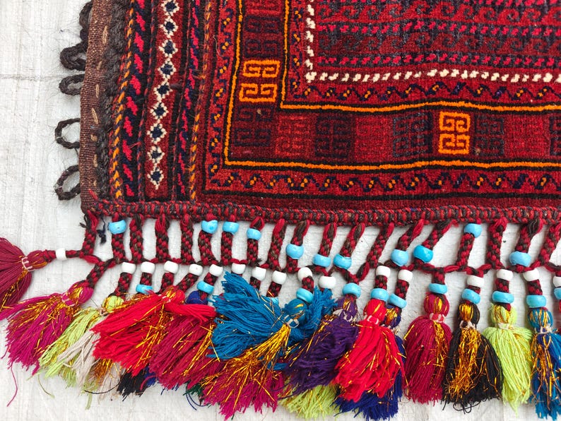 May include: A woven textile featuring a deep red background with intricate geometric patterns in black, orange, and white. The edge is finished with braided fringe adorned with small turquoise and white beads, ending in multi-colored yarn tassels.