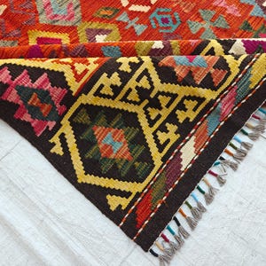 Op de afbeelding: Een geweven kleed met een geometrisch patroon in levendige tinten rood, geel, zwart, blauw en paars. Het kleed is gevouwen en toont een donkerbruine rand met gele ruitmotieven en een franjerand met veelkleurige draadaccenten.
