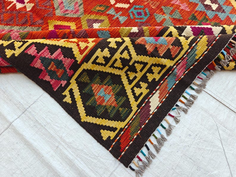 Op de afbeelding: Een geweven kleed met een geometrisch patroon in levendige tinten rood, geel, zwart, blauw en paars. Het kleed is gevouwen en toont een donkerbruine rand met gele ruitmotieven en een franjerand met veelkleurige draadaccenten.