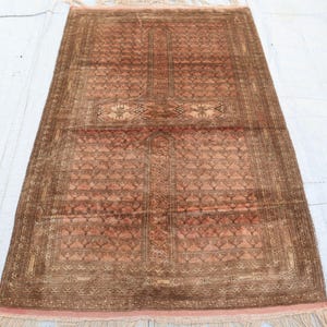May include: A rectangular, hand-woven rug with a central geometric design in shades of brown and peach. The rug features a detailed border and fringe along the edges. The pattern includes repeating shapes and a central medallion.