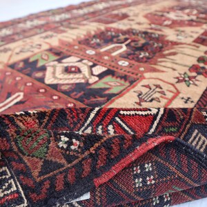 Pu&ograve; includere: Primo piano di un tappeto rosso, nero e crema con intricati motivi geometrici. Il tappeto &egrave; piegato a met&agrave;, rivelando il retro, che &egrave; di colore marrone scuro.