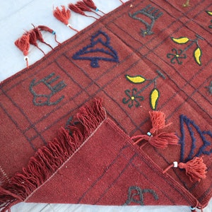May include: A red wool rug with a geometric pattern and fringe. The pattern includes stylized birds, flowers, and leaves in black, blue, and yellow thread.