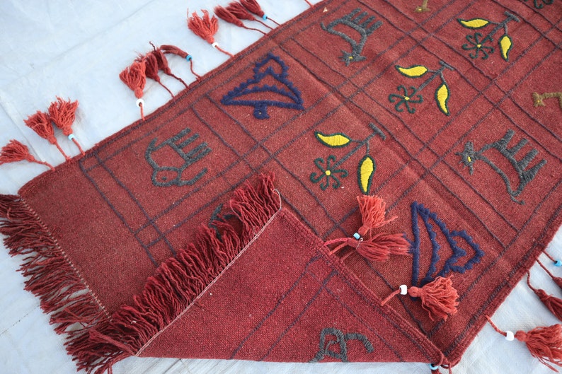 May include: A red wool rug with a geometric pattern and fringe. The pattern includes stylized birds, flowers, and leaves in black, blue, and yellow thread.